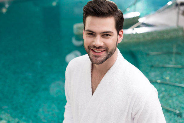 handsome young man in bathrobe smiling at camera while standing in spa center - Foto, immagini