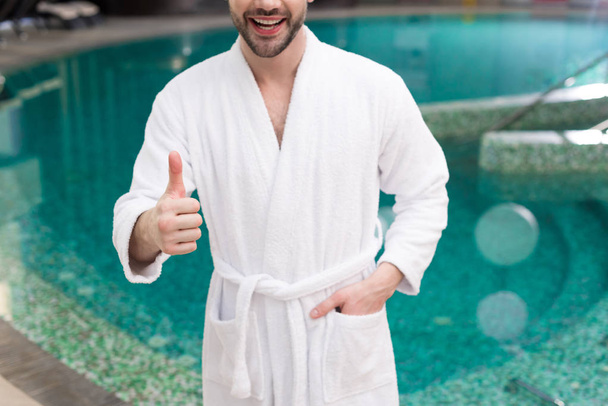 cropped shot of smiling young man in bathrobe showing thumb up in spa center - Foto, Imagem