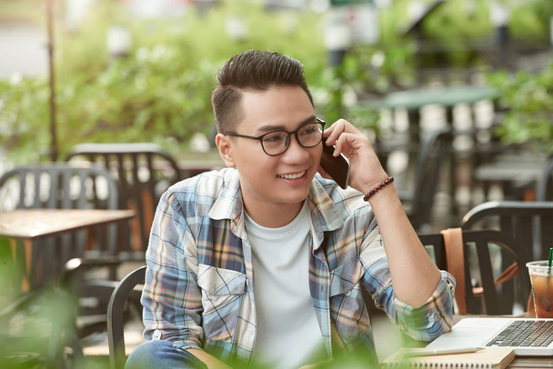 Boldog ázsiai fiatal ember ül a szabadtéri kávézóban, és beszél a telefonon - Fotó, kép