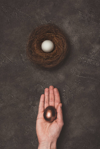cropped view of man holding golden egg in hand, white egg in nest near  - Fotografie, Obrázek