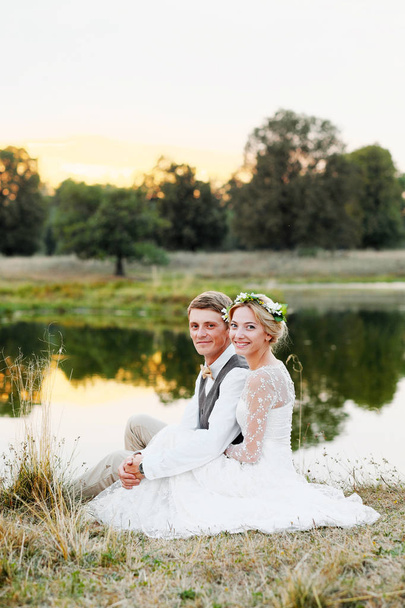 glückliches Paar im Hochzeitskleid vor dem Hintergrund des Sees bei Sonnenuntergang, das Brautpaar in einem weißen Kleid.  - Foto, Bild