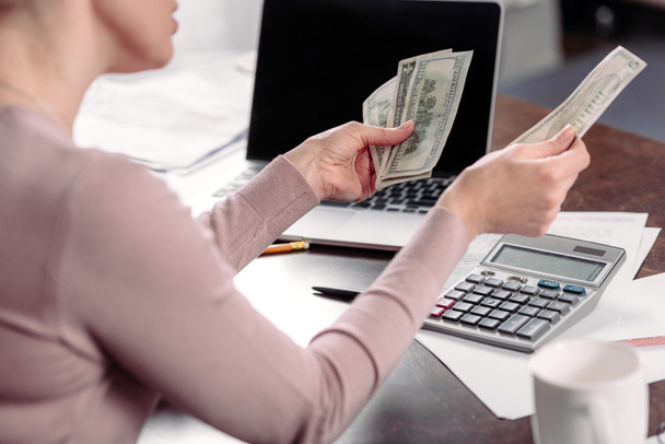 обрезанный снимок женщины, подсчитывающей деньги за столом с ноутбуком
 - Фото, изображение