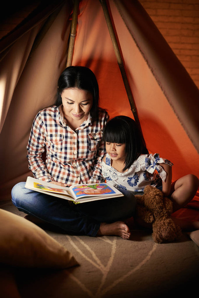 Anya olvasó színes könyv a lányának, amikor ők ülnek egy sátorban - Fotó, kép