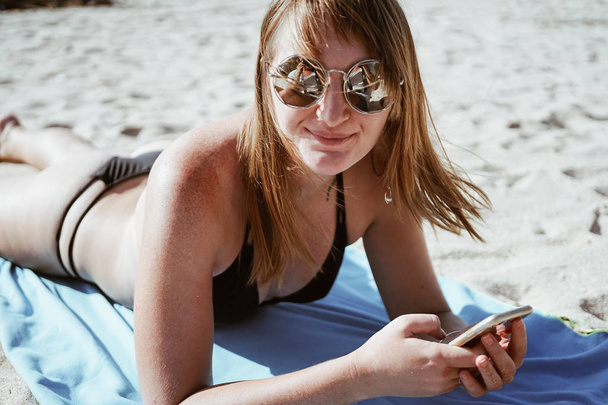 Porträt eines rothaarigen Mädchens mit runder Spiegelsonnenbrille. das Mädchen liegt im Sand am Meer. - Foto, Bild
