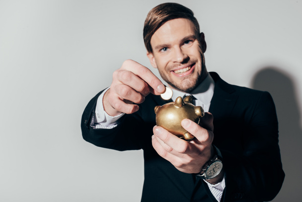 Молодой уверенный бизнесмен в костюме кладет монету в золотую копилку на белом фоне
 - Фото, изображение