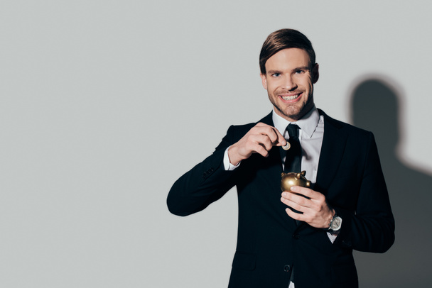 Smiling businessman in suit putting coin in golden piggy bank on white background - Fotografie, Obrázek
