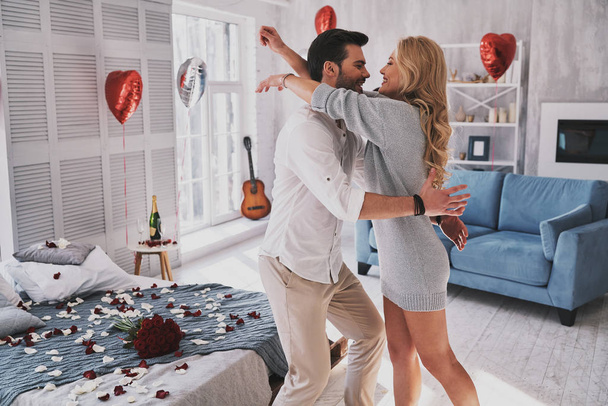 Totally in love beautiful couple embracing in bedroom full of balloons and rose petals, celebrating valentines day and engagement - Photo, Image