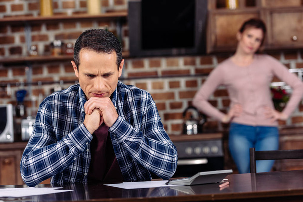 selective focus of pensive man sitting at table and wife standing behind at home, financial problems concept - Photo, Image