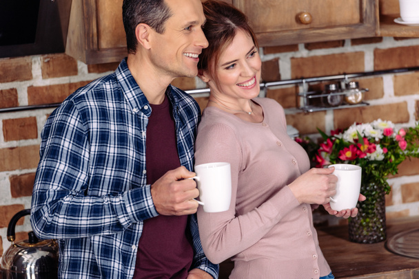 πορτρέτο του χαμογελώντας γυναίκα και άντρα με κούπες καφέ στην κουζίνα στο σπίτι - Φωτογραφία, εικόνα
