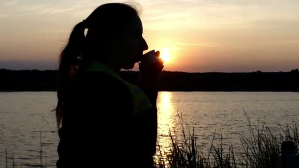 Miła kobieta siedzi na brzegu jeziora, picie herbaty z termos - Materiał filmowy, wideo