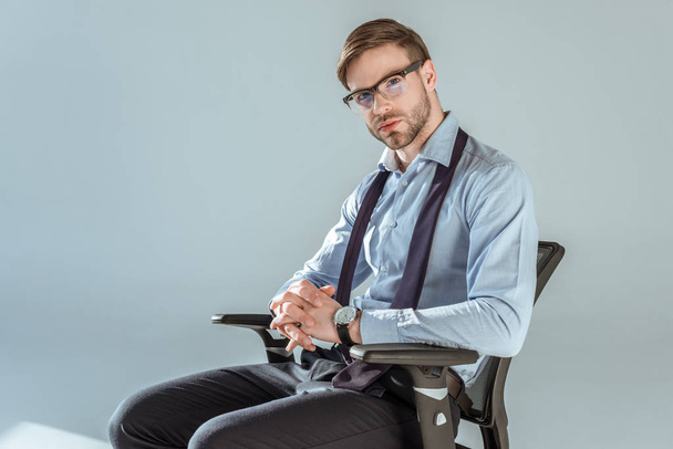 Young confident businessman sitting on chair with hands clenched,  isolated on grey - Photo, Image