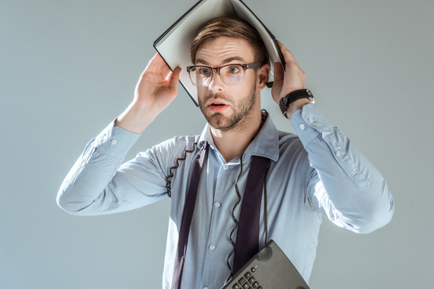 Jonge verrast zakenman holding laptop op zijn hoofd geïsoleerd op grijs - Foto, afbeelding