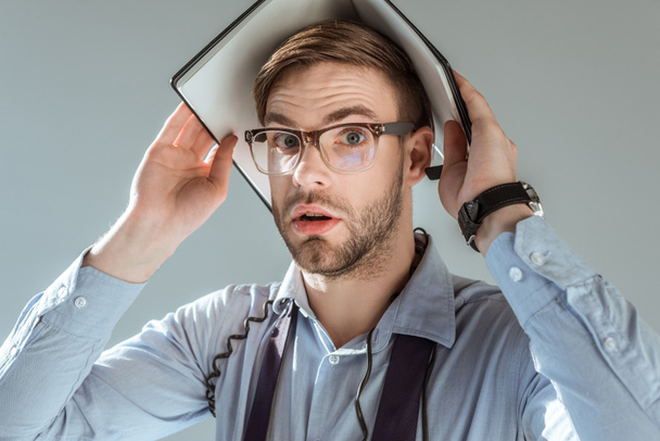 Empresário confuso segurando caderno em sua cabeça isolado em cinza
 - Foto, Imagem