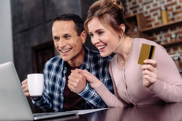 happy couple with credit card and cup of coffee looking at laptop screen at home - Foto, imagen