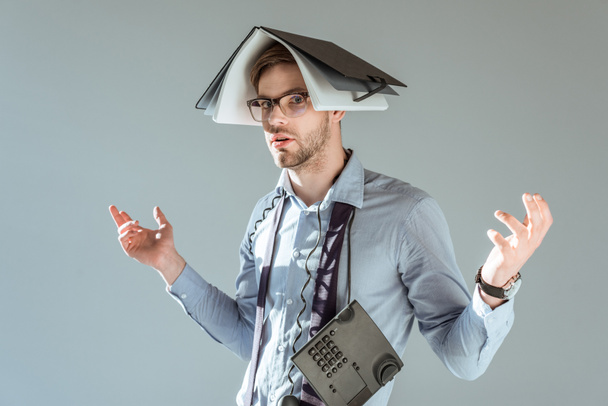 Young confused businessman holds notebook on his head and phone on neck isolated on grey - Foto, Imagen