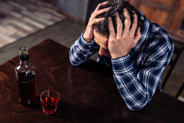wysoki kąt widzenia pijany mężczyzna siedzący przy stole z butelkę i kieliszek alkoholu w domu - Zdjęcie, obraz