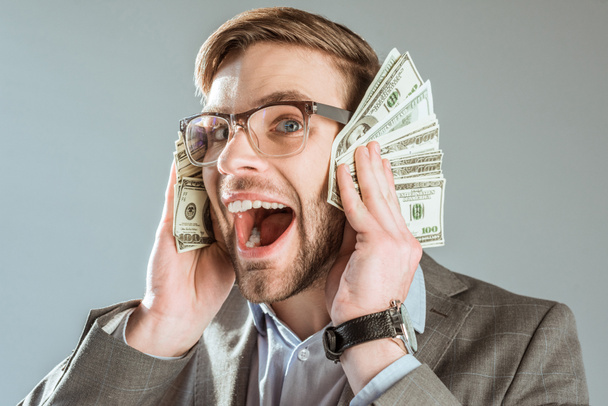 rijke gelukkig zakenman holding dollarbiljetten in de buurt van zijn gezicht geïsoleerd op grijs - Foto, afbeelding