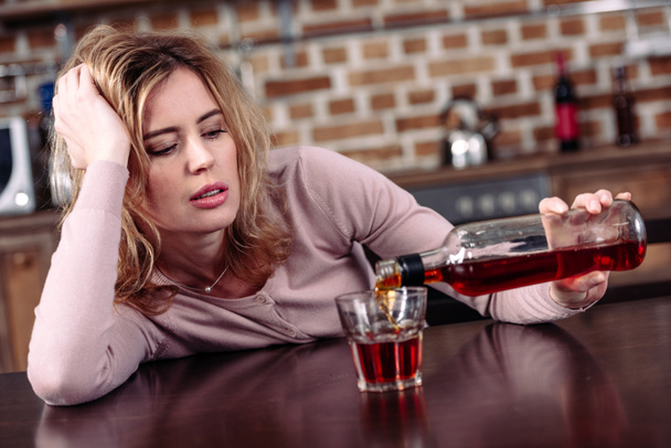 γυναίκα ρίχνει αλκοόλ στο γυαλί, ενώ κάθεται στο τραπέζι στην κουζίνα - Φωτογραφία, εικόνα