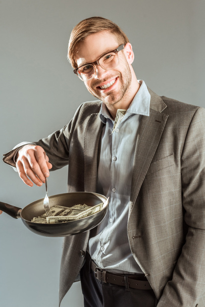 Young businessman frying dollar bills in pan isolated on grey - Photo, Image