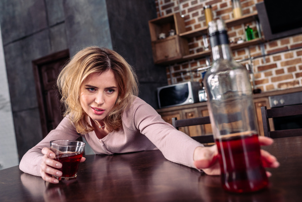 enfoque selectivo de la mujer borracha tomando botella de alcohol mientras está sentada en la mesa en casa
 - Foto, Imagen