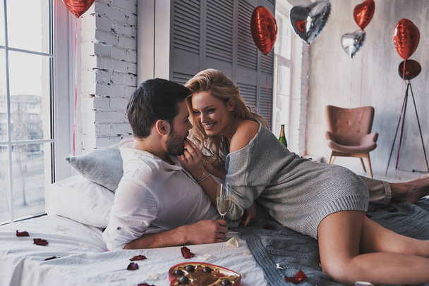 Beautiful young couple lying on bed at home, woman feeding man with chocolate - Φωτογραφία, εικόνα