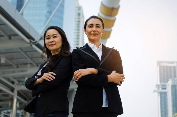 two smiling business asian and caucasian woman in formally dressed looking into camera with her arms crossed standing in modern city, confident, team leader, successful and entrepreneurship concept - Zdjęcie, obraz