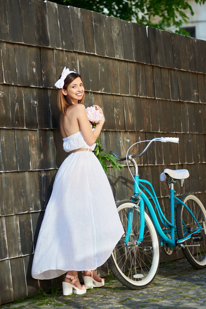 happy young beautiful woman holding bunch of peonies posing elegantly near bicycle outdoors  - Photo, Image