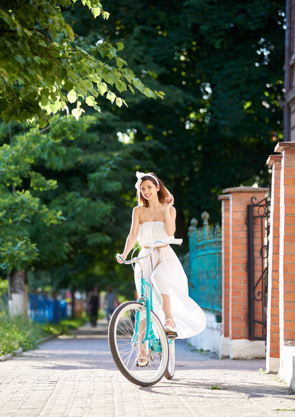 gorgeous cheerful young woman in long white dress cycling on bicycle in park   - Фото, изображение
