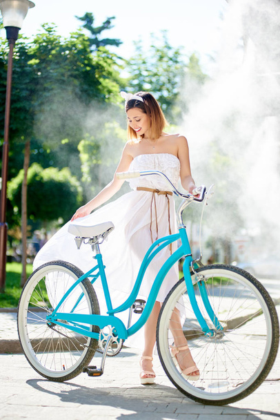 蒸し暑い午後の夏カフェの霧システムの下でブルーのレトロな自転車と光のドレスを着たかわいい女性 - 写真・画像