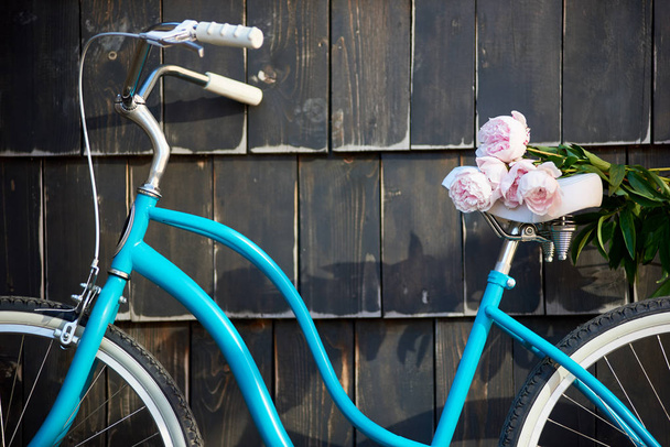 Close-up of blue vintage bicycle with bouquet of gently pink peonies on dark wall background - Фото, изображение
