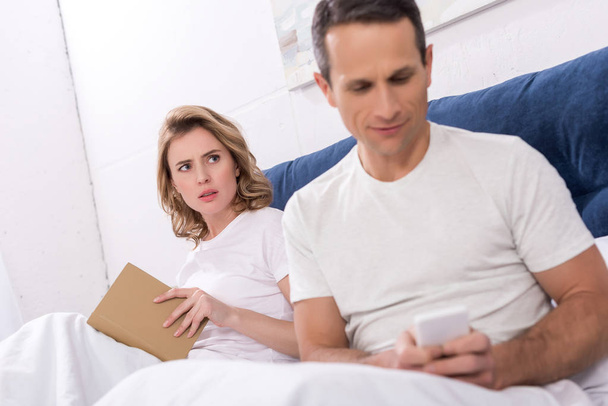 Selektywny fokus człowieka za pomocą smartfona, natomiast żona czytania książki w łóżku - Zdjęcie, obraz