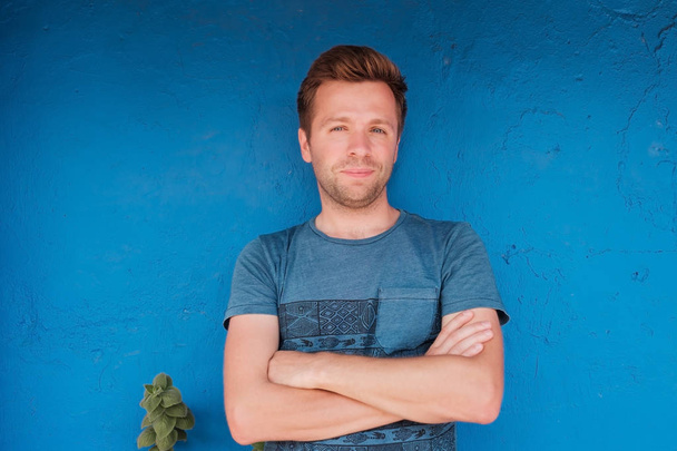 Portrait of smiling young caucasian man standing near blue wall with freen leaf - Photo, Image