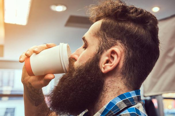 Крупным планом портрет красивого стильного бородатого мужчину с татуировкой на руке, одетый во фланелевую рубашку пьет кофе в парикмахерской
. - Фото, изображение