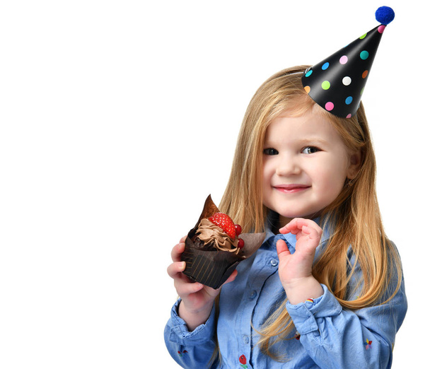 Three years girl kid eat sweet chocolate cake muffin with fruits celebrating in birthday cap isolated  - Foto, Bild