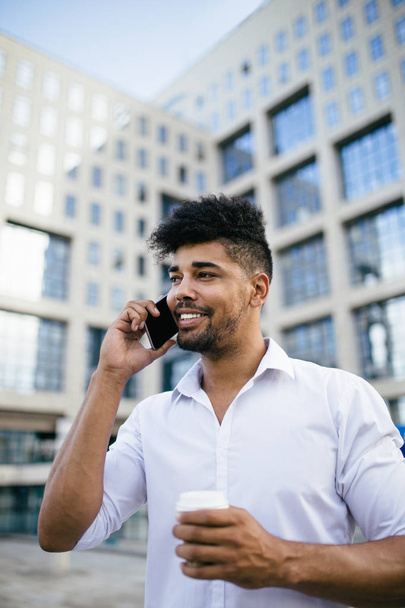 Young handsome Afro American man standing in front of huge modern business building smiling and talking on cell phone - Foto, Imagem