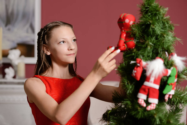 het meisje in de rode jurk naast de witte haard, decoreren een kerstboom met speelgoed - Foto, afbeelding