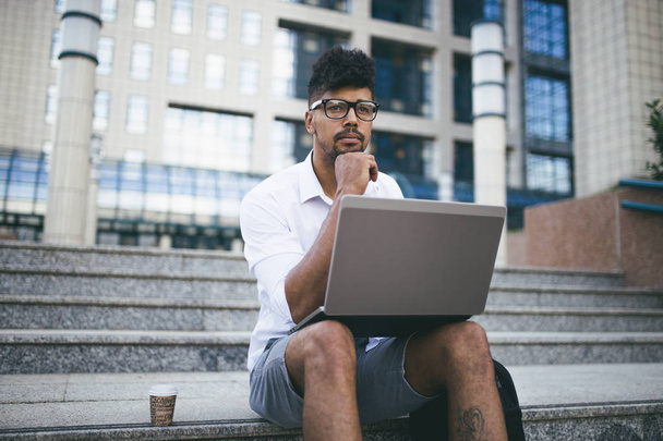 Young attractive afro American businessman sitting on stairs in front of huge modern building and doing some work on laptop - Foto, immagini