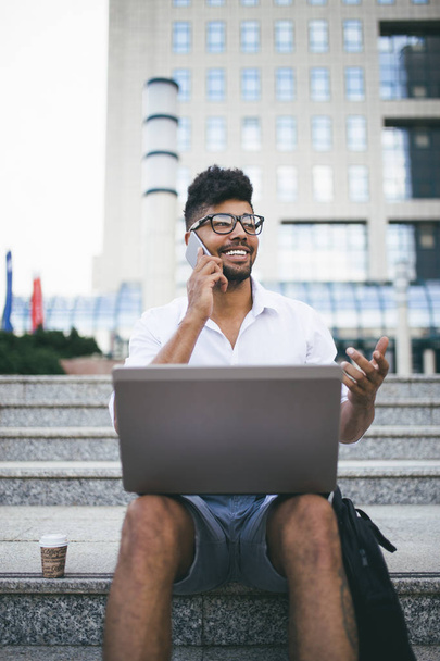 Young attractive afro American businessman sitting on stairs in front of huge modern building and doing some work on laptop - Foto, Imagen