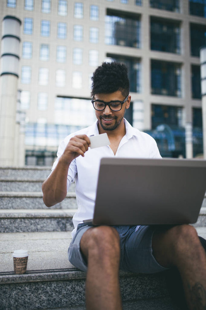 Young attractive afro American businessman sitting on stairs in front of huge modern building and doing some work on laptop - Φωτογραφία, εικόνα