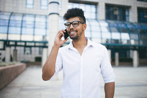 Young handsome Afro American man standing in front of huge modern business building smiling and talking on cell phone - Fotoğraf, Görsel