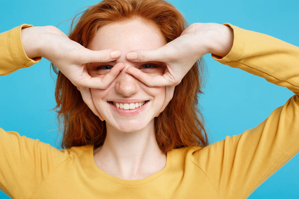 Fun and People Concept - Headshot Portrait of happy ginger red hair girl with freckles smiling and making finger glasses. Pastel blue background. Copy Space. - Fotografie, Obrázek