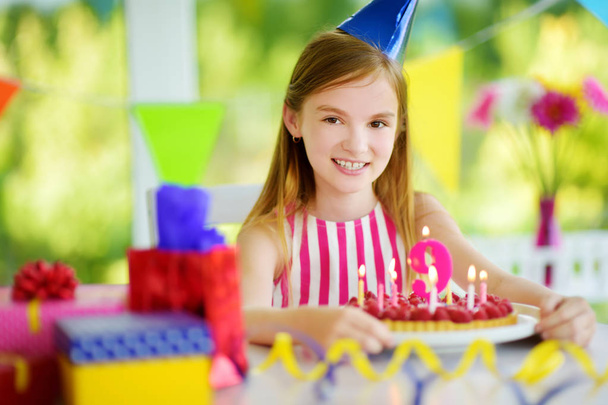 entzückendes Mädchen, das zu Hause Geburtstagsparty feiert und Kerzen auf Geburtstagstorte pustet. Kindergeburtstag mit bunten Dekorationen, Geschenken und Fahnen. - Foto, Bild