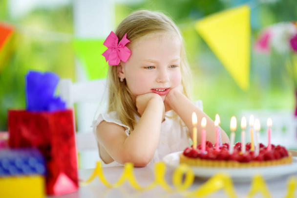 Adorable girl having birthday party at home, blowing candles on birthday cake. Kids birthday party with colorful decorations, gifts and banners. - Photo, image