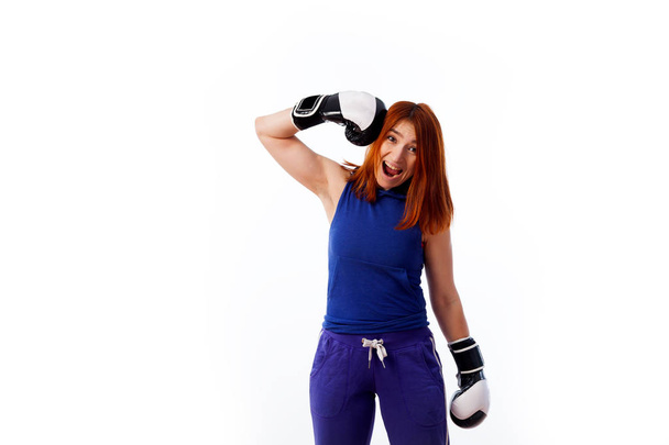 young red-haired woman in sportswear and black and white boxing gloves smiles and weakly strikes herself with two gloves on the head on a white isolated background - Photo, Image