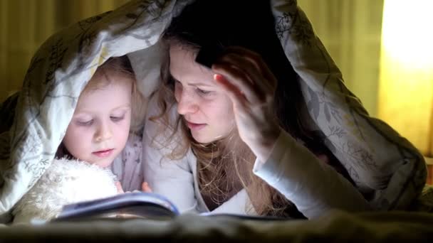 母と娘のベッドのカバーの下で本を読むと懐中電灯を持って笑みを浮かべてください。. - 映像、動画