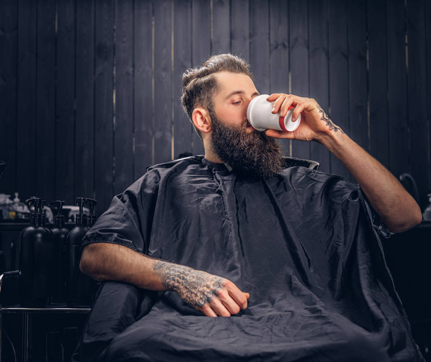 Handsome bearded man with a tattoo on his arms drinks coffee before hair wash in a hairdressers salon. - Photo, Image