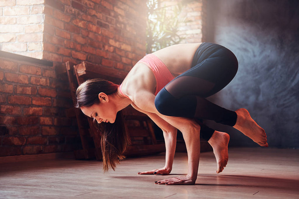 attraktive Frau mittleren Alters balanciert auf Händen und praktiziert Yoga in einem Raum mit Loft-Interieur. - Foto, Bild