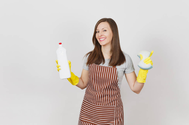 Young smiling housewife in yellow gloves, striped apron isolated on white background. Attractive woman holding bottle with cleaner liquid for washing and cleaning, sponge. Copy space for advertisement - Zdjęcie, obraz