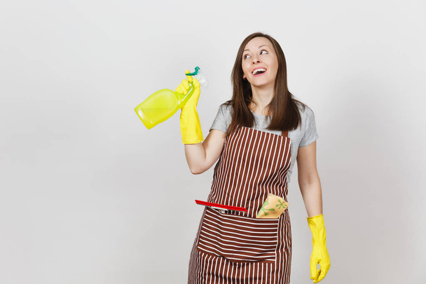 Young smiling housewife in yellow gloves, striped apron, cleaning rag, squeegee in pocket isolated on white background. Housekeeper woman holding spray bottle with cleaner liquid. Bottle copy space. - Foto, imagen
