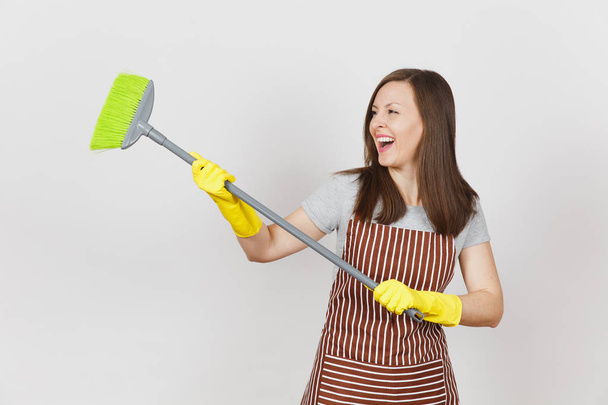 junge lächelnde Hausfrau in gestreifter Schürze, gelbe Handschuhe isoliert auf weißem Hintergrund. Spaß Haushälterin Putzfrau halten und kehren mit Besen. Kopierfläche für Werbefläche - Foto, Bild